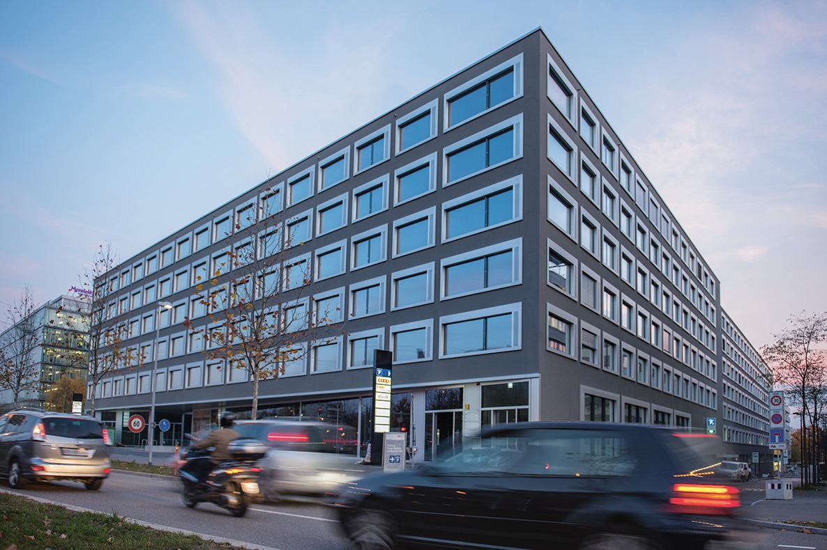 A modern, multi-story office building with a sleek, glass-and-steel facade. The building has numerous windows and appears to be located on a busy street with traffic passing by.