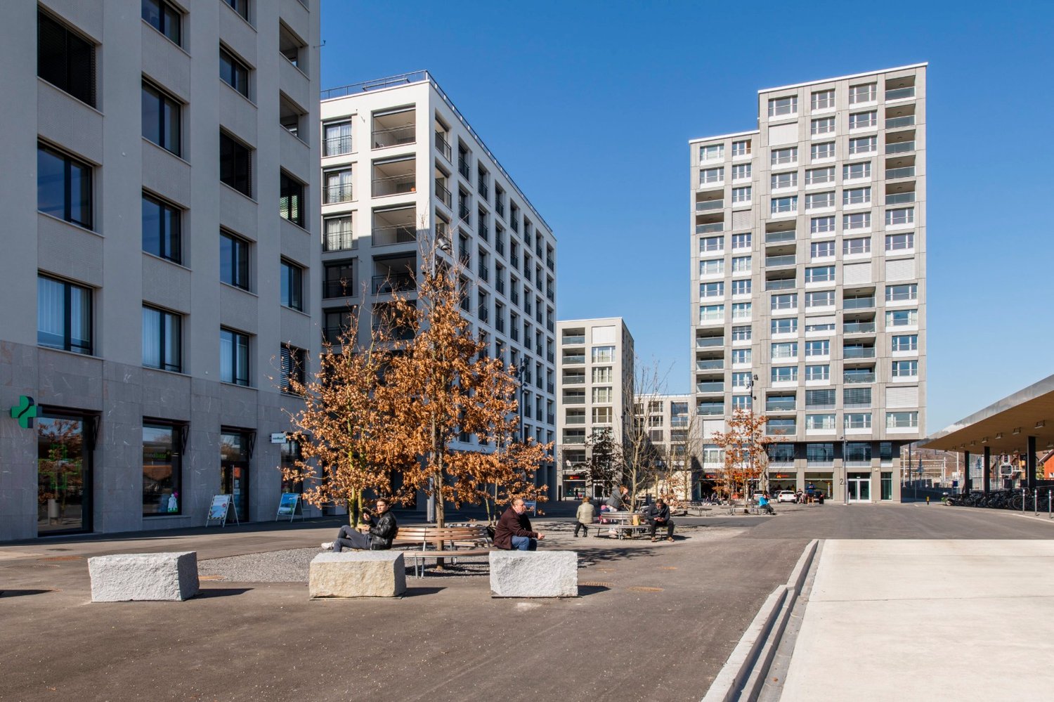 The image shows a modern apartment building complex with several high-rise buildings. The buildings have balconies and large windows. There is a public plaza in the foreground with benches and trees, where people are sitting and relaxing.