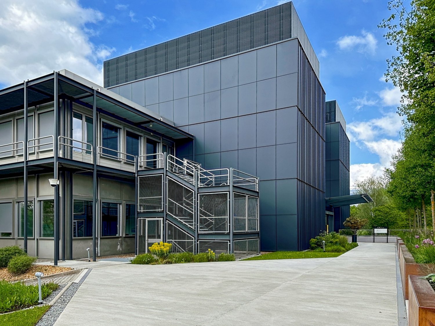 modern building, gray exterior, large windows, entrance, concrete pathway, shrubs, trees