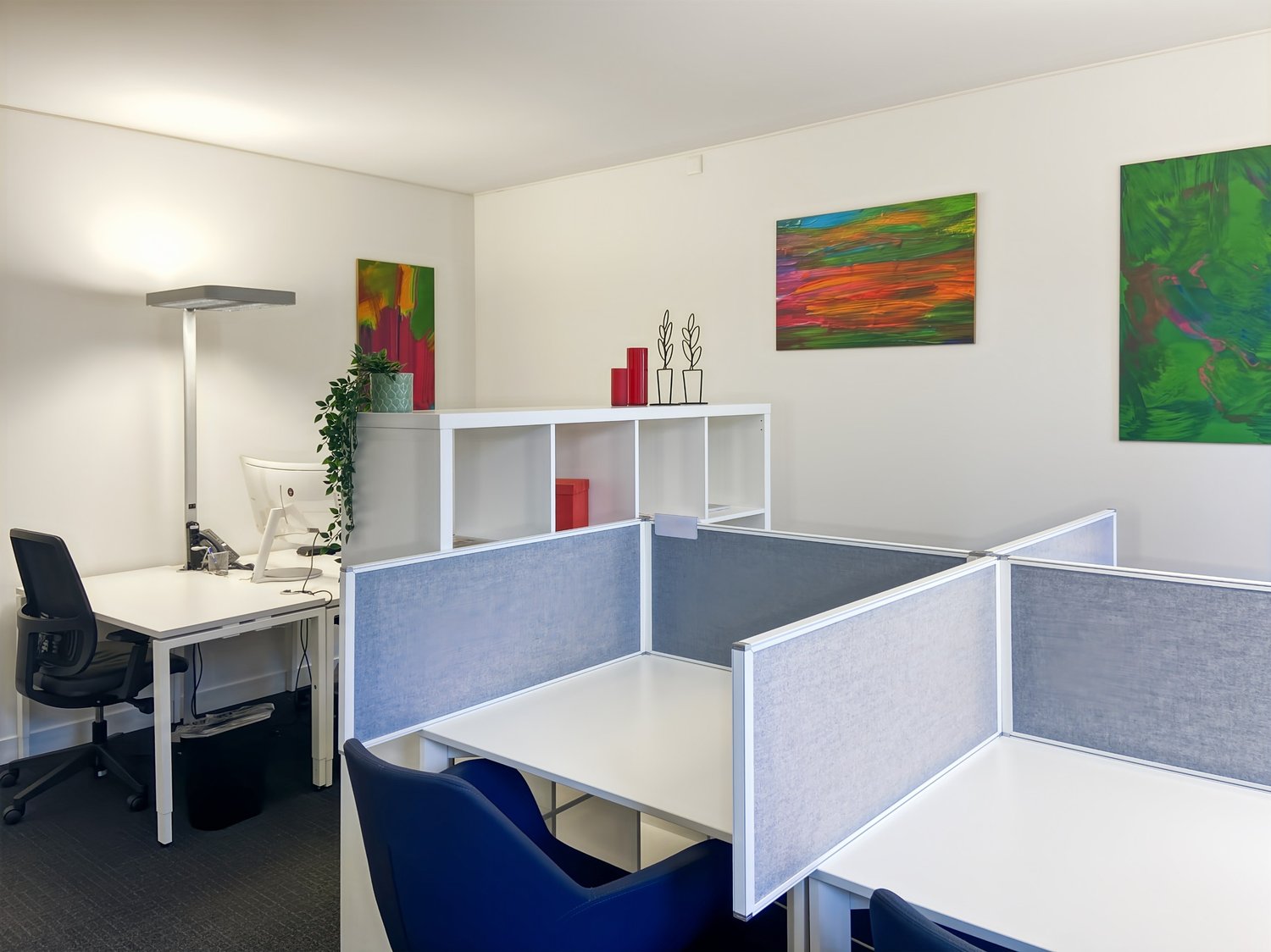 The image shows an office space with a desk, computer, and office chair. There are also cubicle-style partitions separating different workspaces, and colorful artwork on the walls. The space appears to be well-lit and organized.