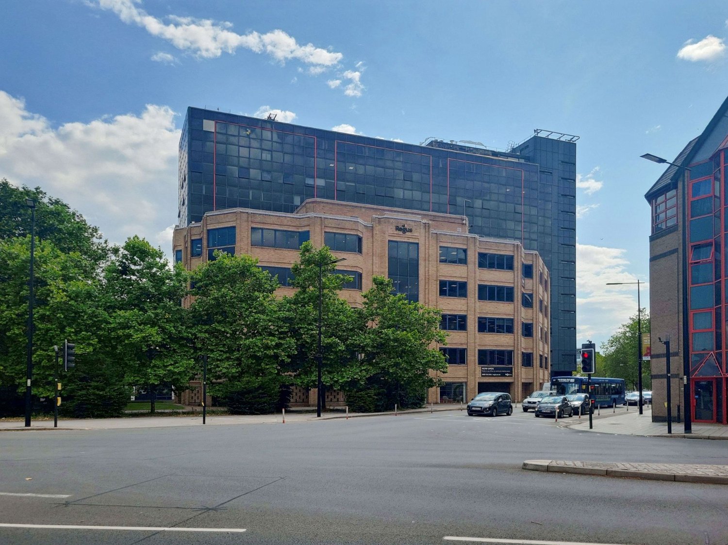 Commercial building with two sections, lower brown brick section and taller glass section, multiple cars parked on street, several trees, traffic lights, and buses on road