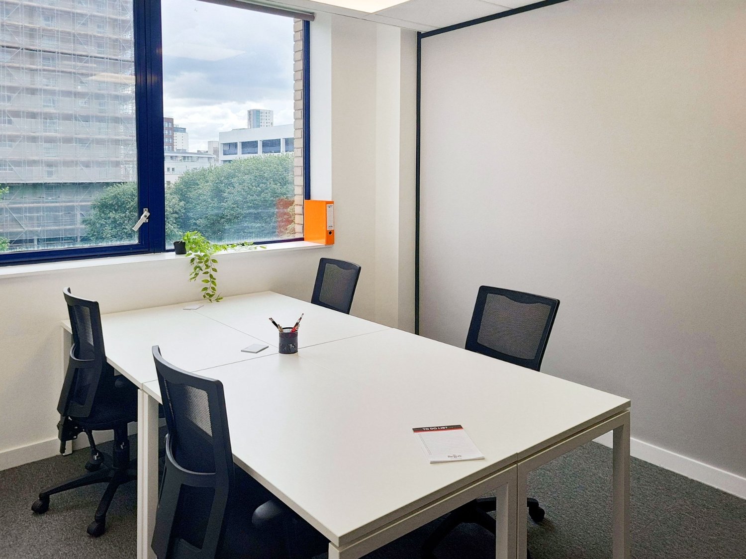 Modern office meeting room with a long white table, four black office chairs, white walls, and large windows with a view of the outside. A small potted plant and a small orange file are on the window sill. A notepad and pens are on the table.
