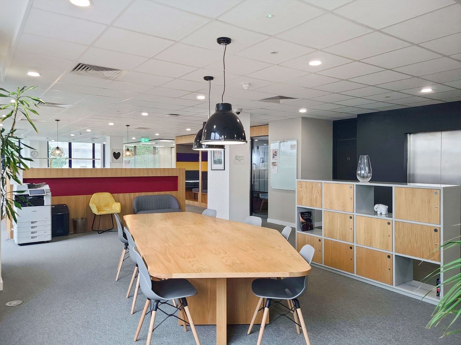 large table, gray and yellow chairs, gray and white shelves, photo frame on wall, hanging lamps, printer, trash can, clock
