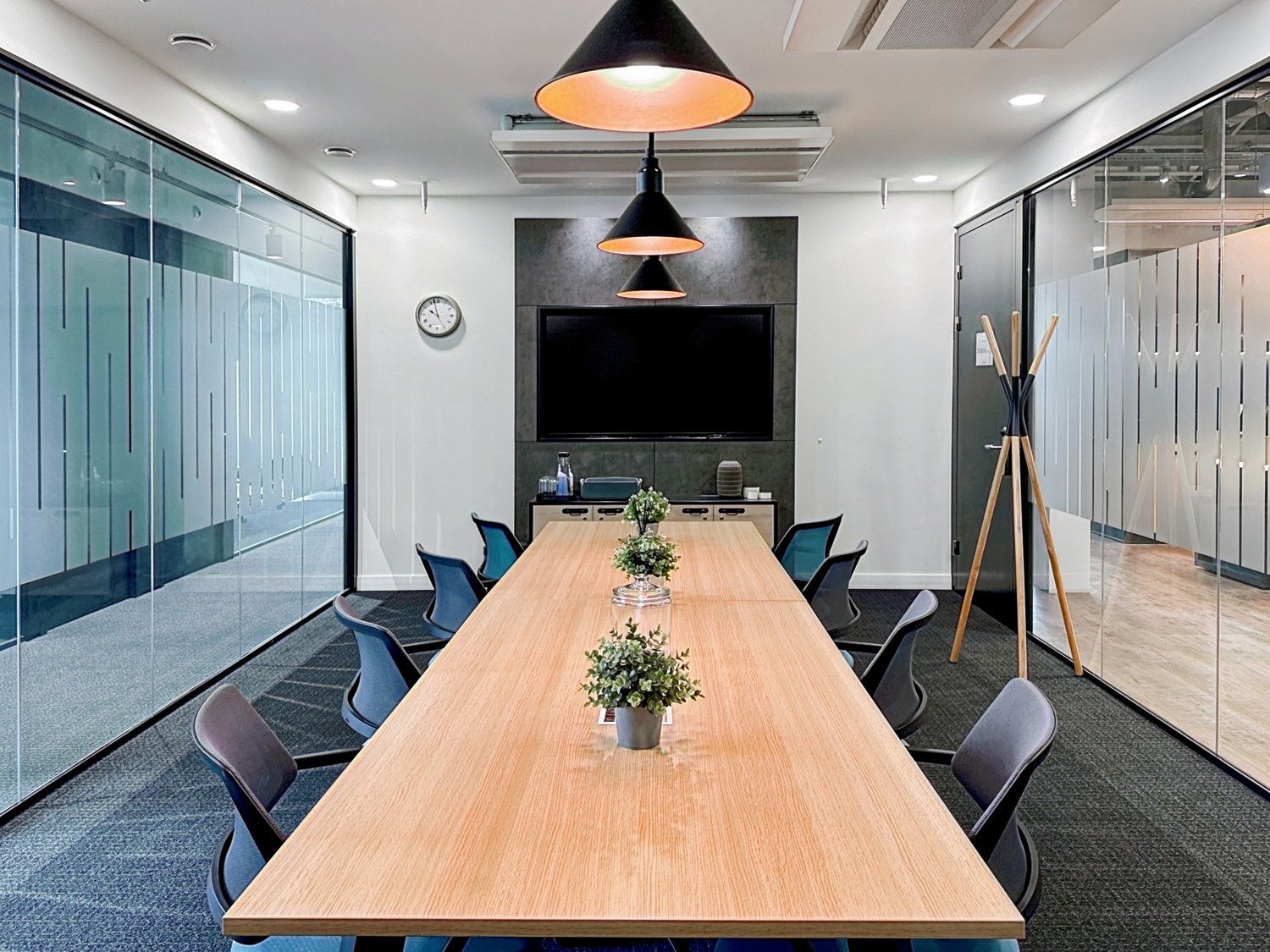 Long wooden table, blue chairs, white wall, black TV, clock, potted plants