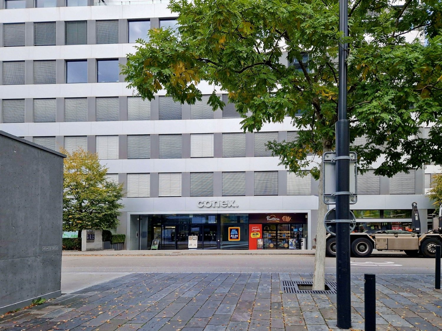 Office building with glass facades, glass doors, and metal columns, surrounded by trees and parked vehicles