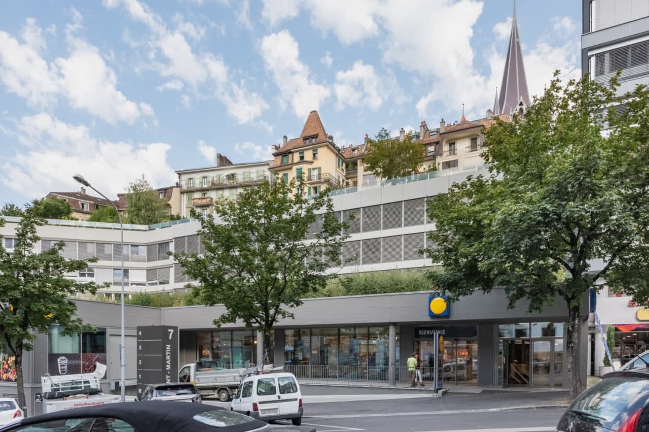 Multi-story building with a mix of residential and commercial spaces, including a supermarket. The building has a distinctive architectural style with a spire-like structure on top. There are balconies visible on some of the upper floors.