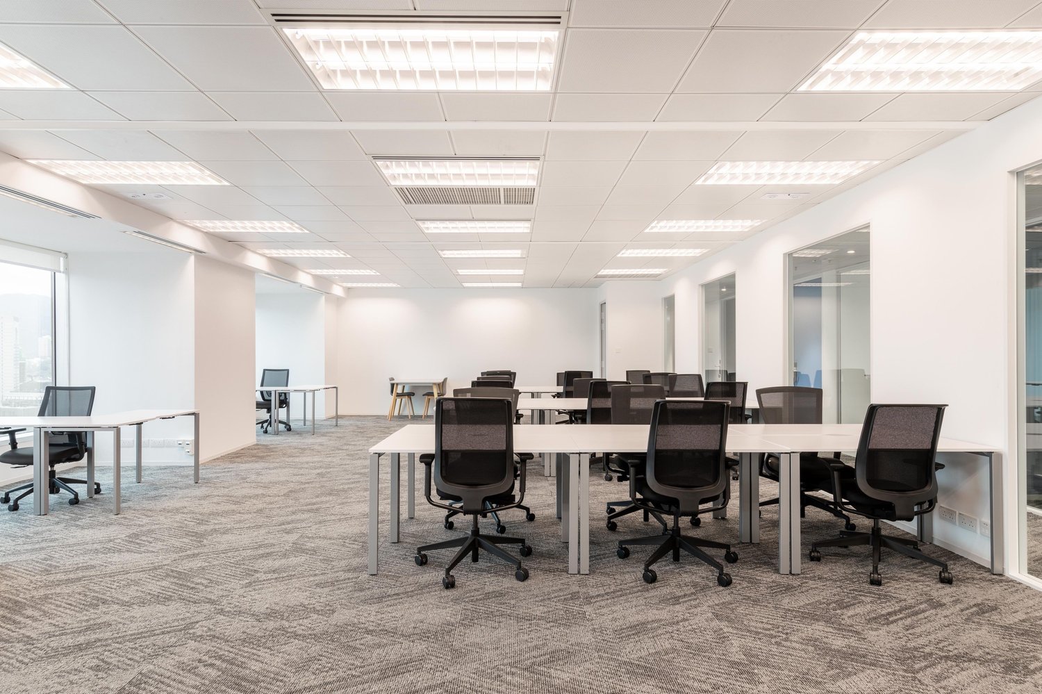 Large open office space with multiple workstations, desks, and chairs. The room has a modern and minimalist design with white walls, a tiled floor, and recessed lighting. There are glass partitions and doors visible, indicating separate office areas or me