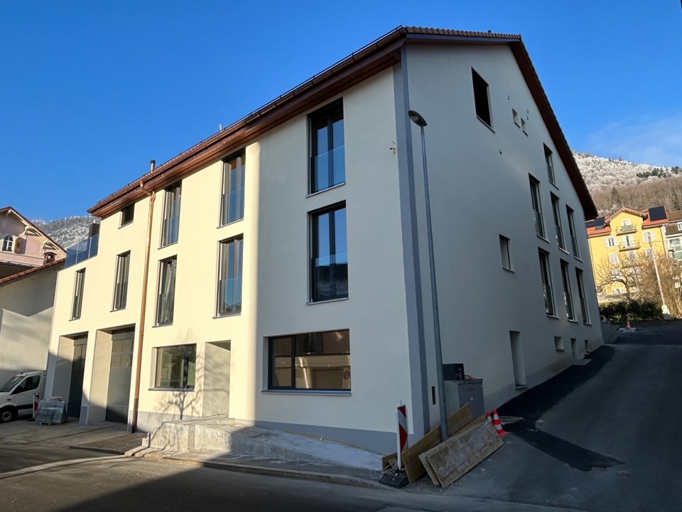 Apartment building with several units, white walls, brown roof, large windows, ground floor entrance, white van parked in front.