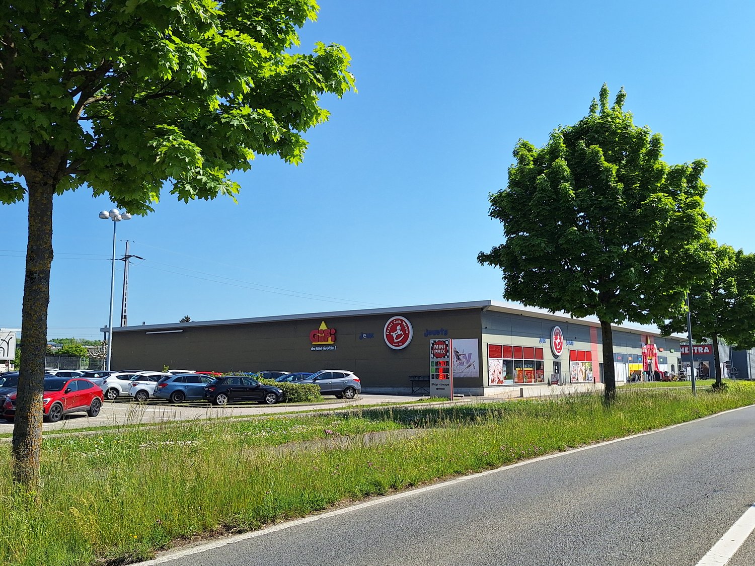 A large commercial building with a parking lot in front, surrounded by trees and greenery. The building has a black exterior with various signs and logos, indicating it is likely a retail or shopping center.