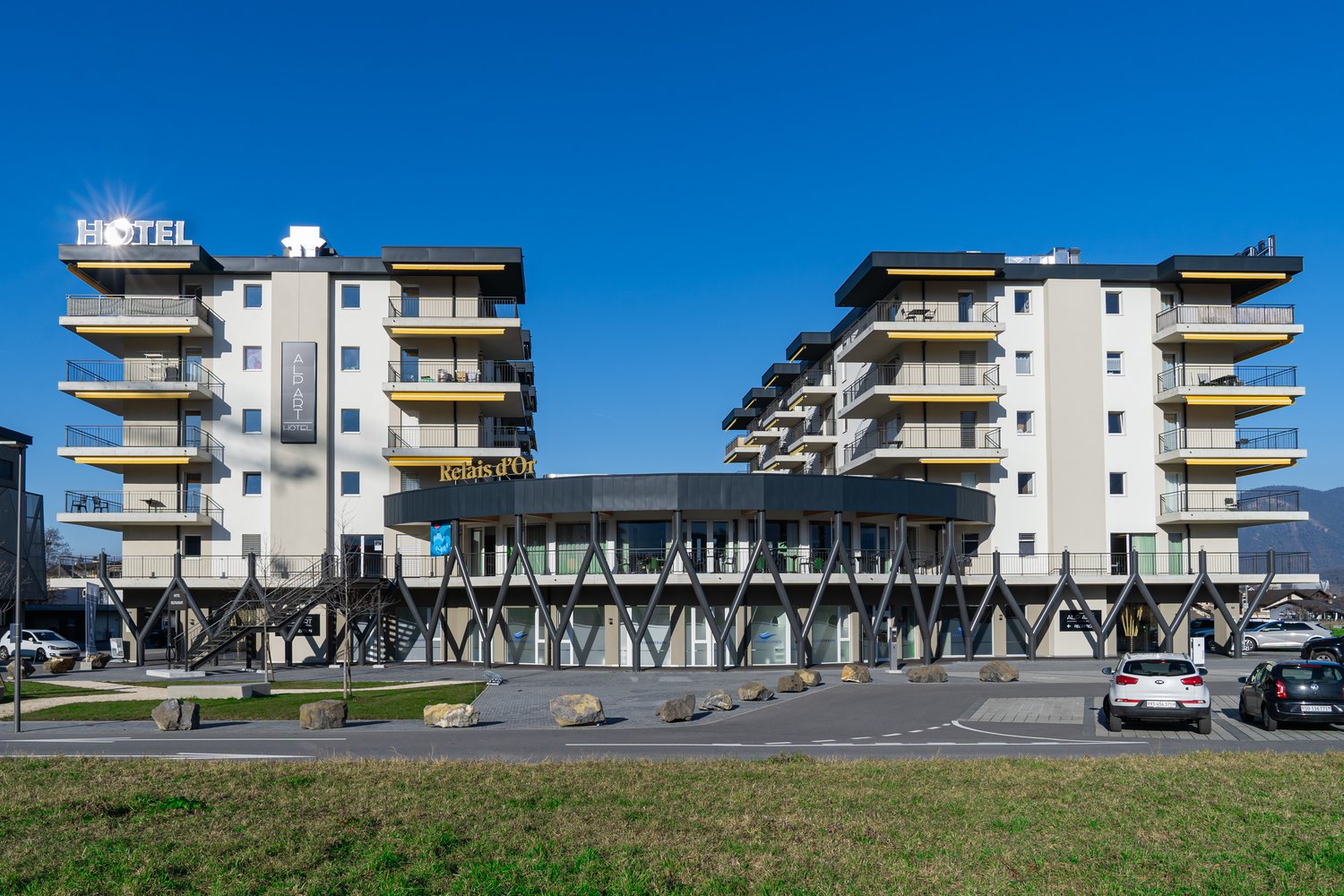 Modern multi-story hotel building with balconies, glass entrance, and parking area in front