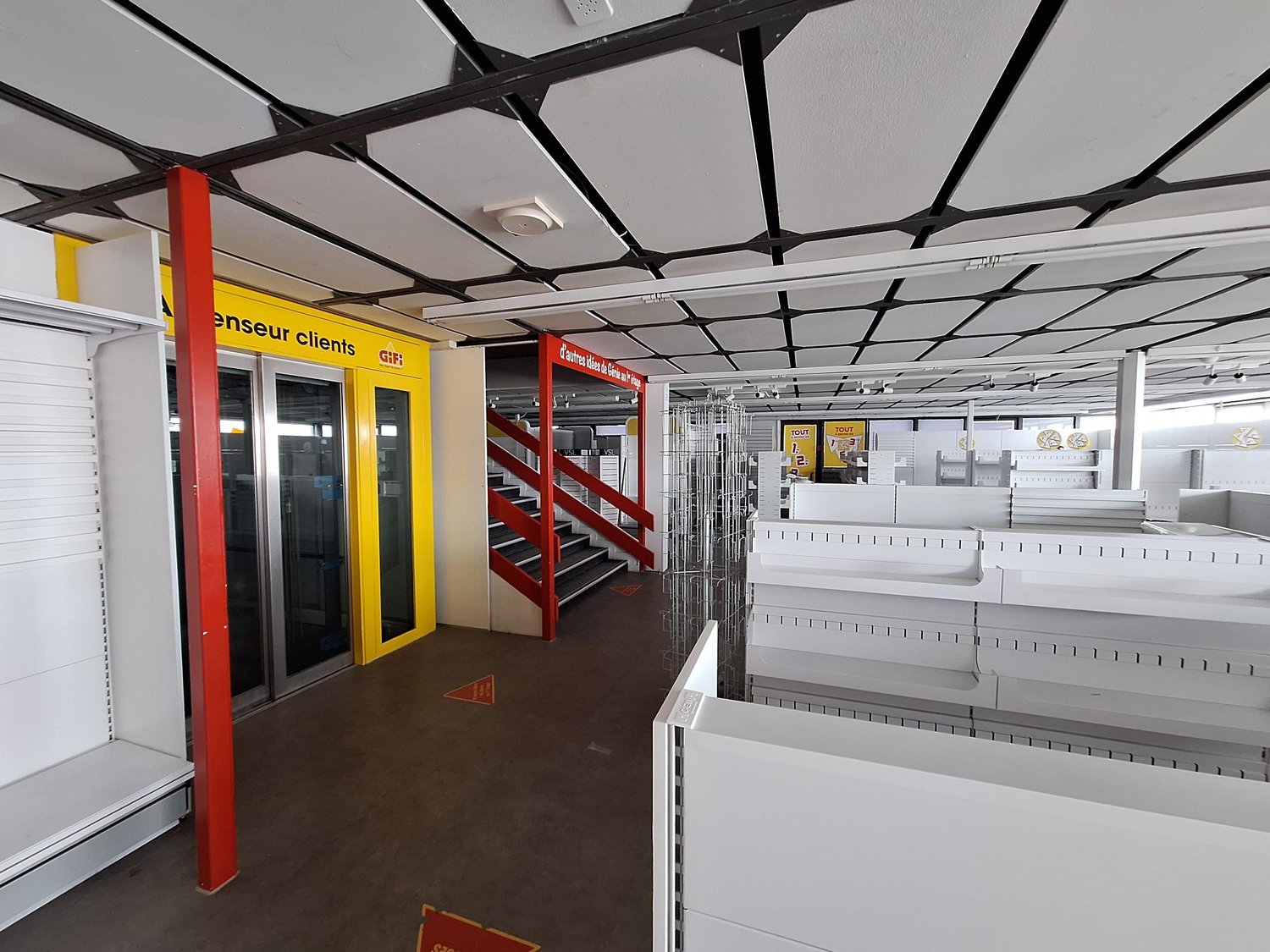 Empty room with white shelves, red and white signs, glass doors, and a staircase