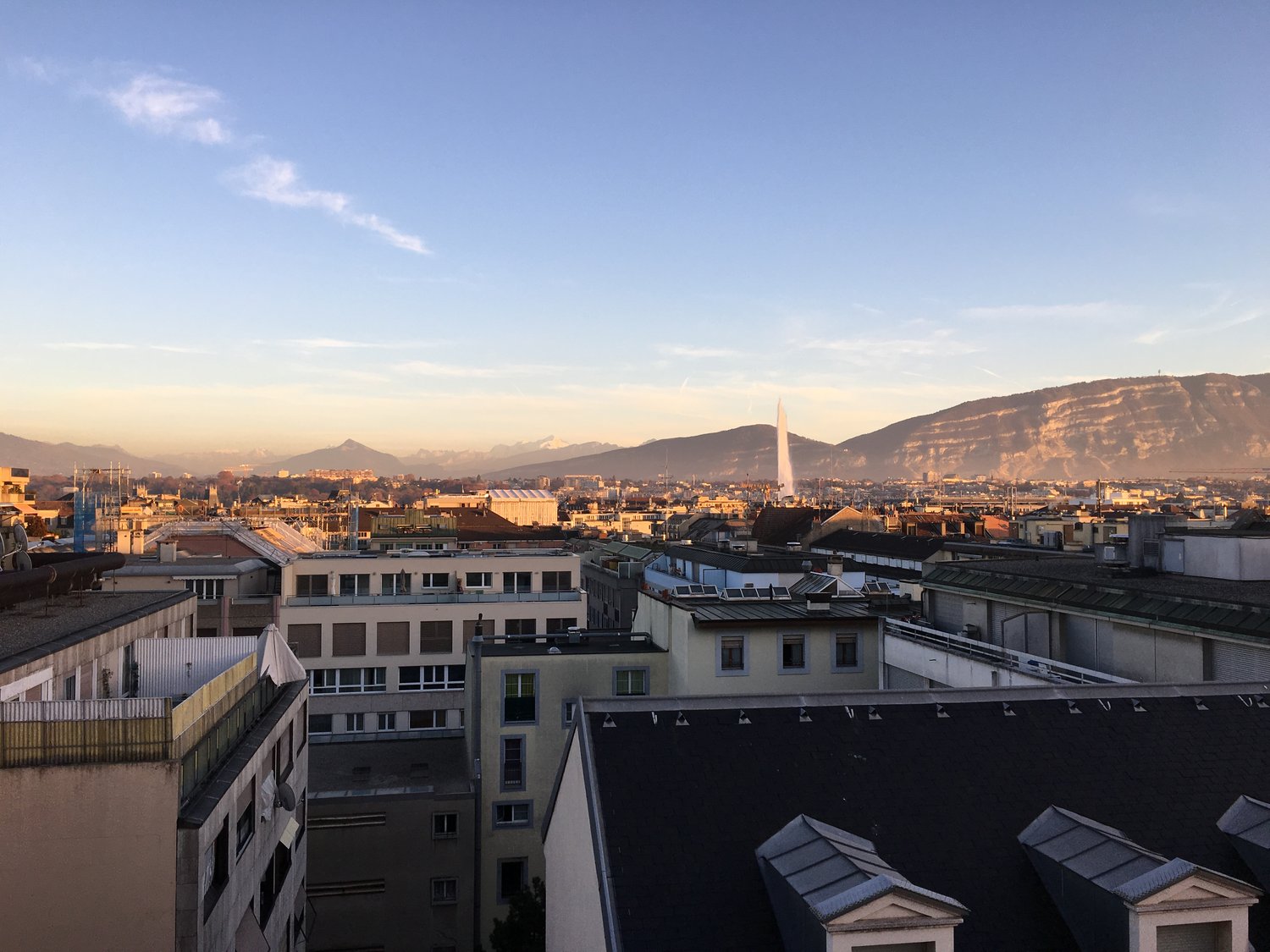 Cityscape with multiple buildings, mountains in the background, and a fountain in the distance