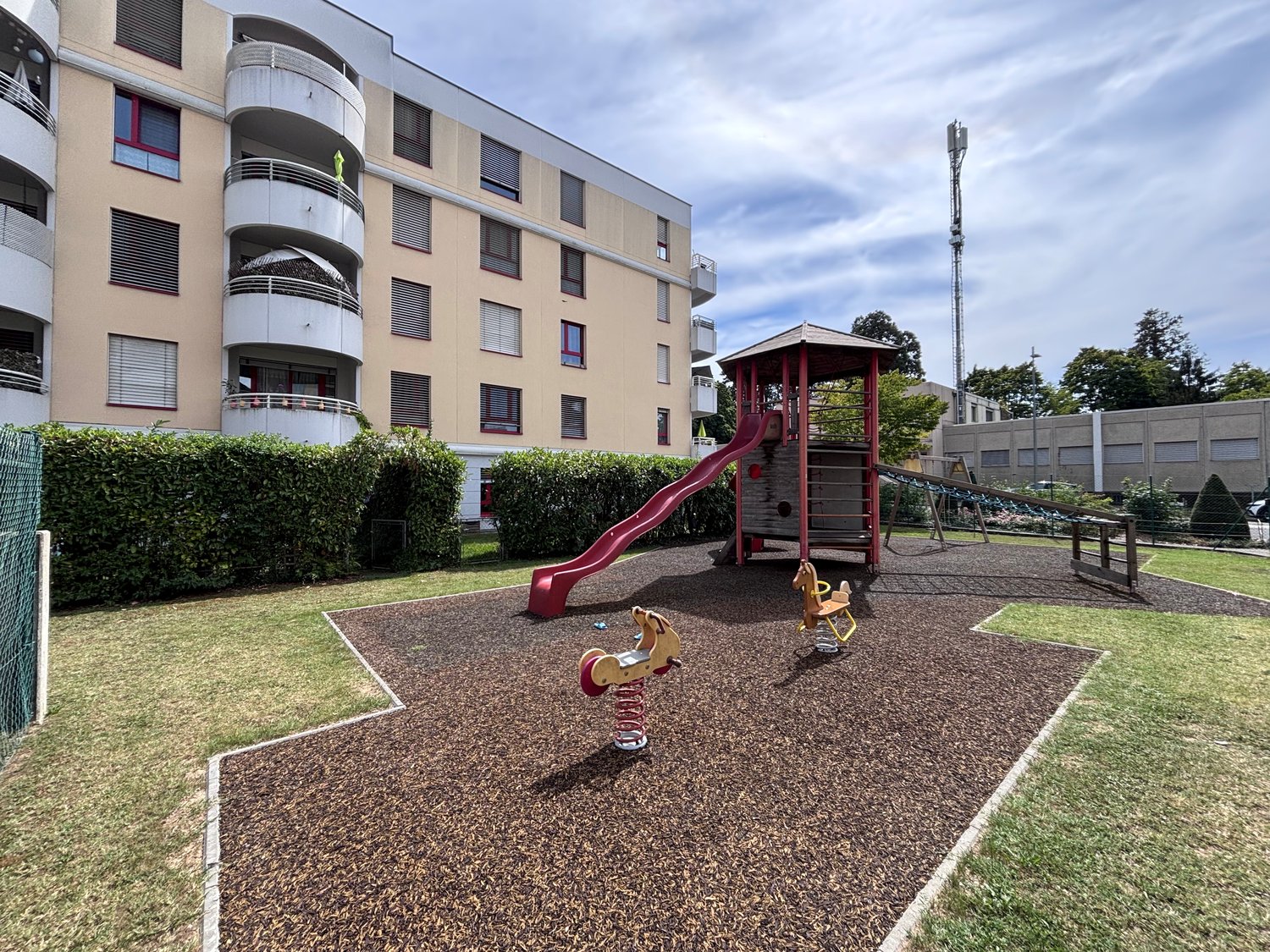 Playground with slide, climbing structure, and horsey ride toys, bordered by bushes, adjacent to a building with balconies.