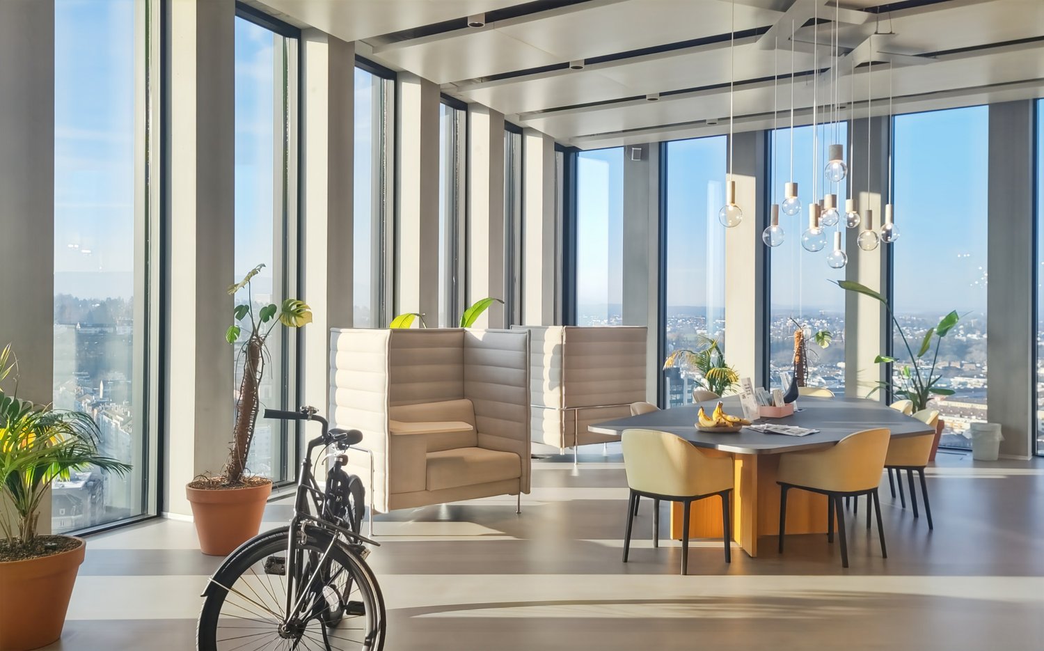 Spacious living room with floor-to-ceiling windows offering a panoramic view of the city skyline. The room features comfortable beige armchairs, a dining table with chairs, and a bicycle. Decorative plants and pendant lighting add to the modern and stylis