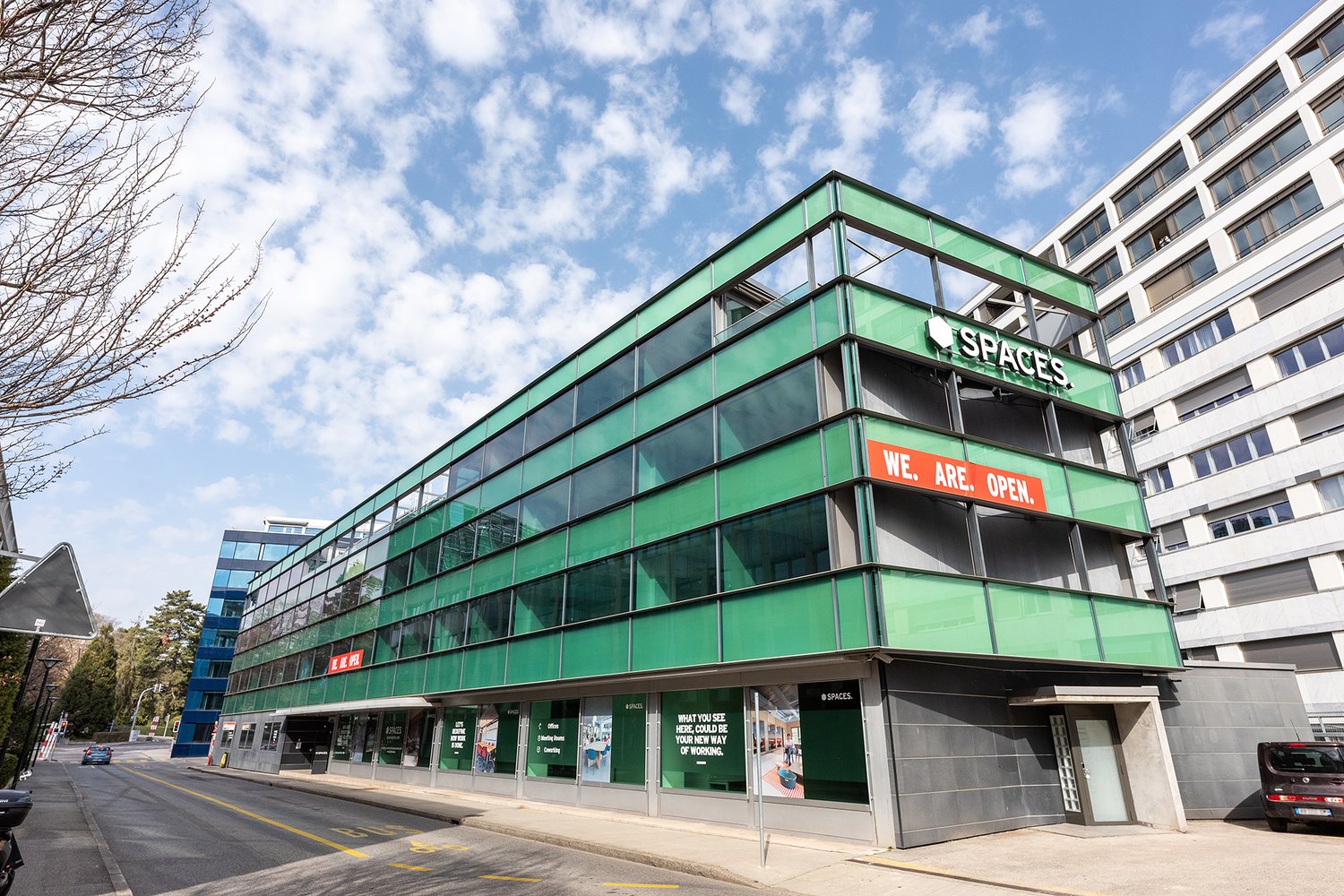 A multi-story commercial building with a green glass facade and the 'Spaces' logo. The building has large windows and a 'We. Are. Open.' sign displayed prominently. There are parking spaces visible in front of the building.