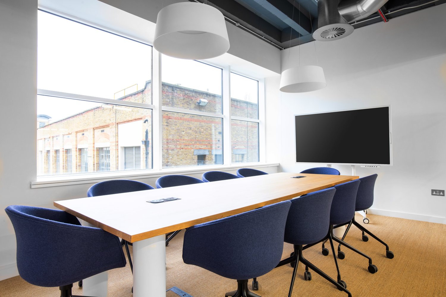 Large meeting room with wooden table, blue office chairs, large windows, and a TV screen on the wall