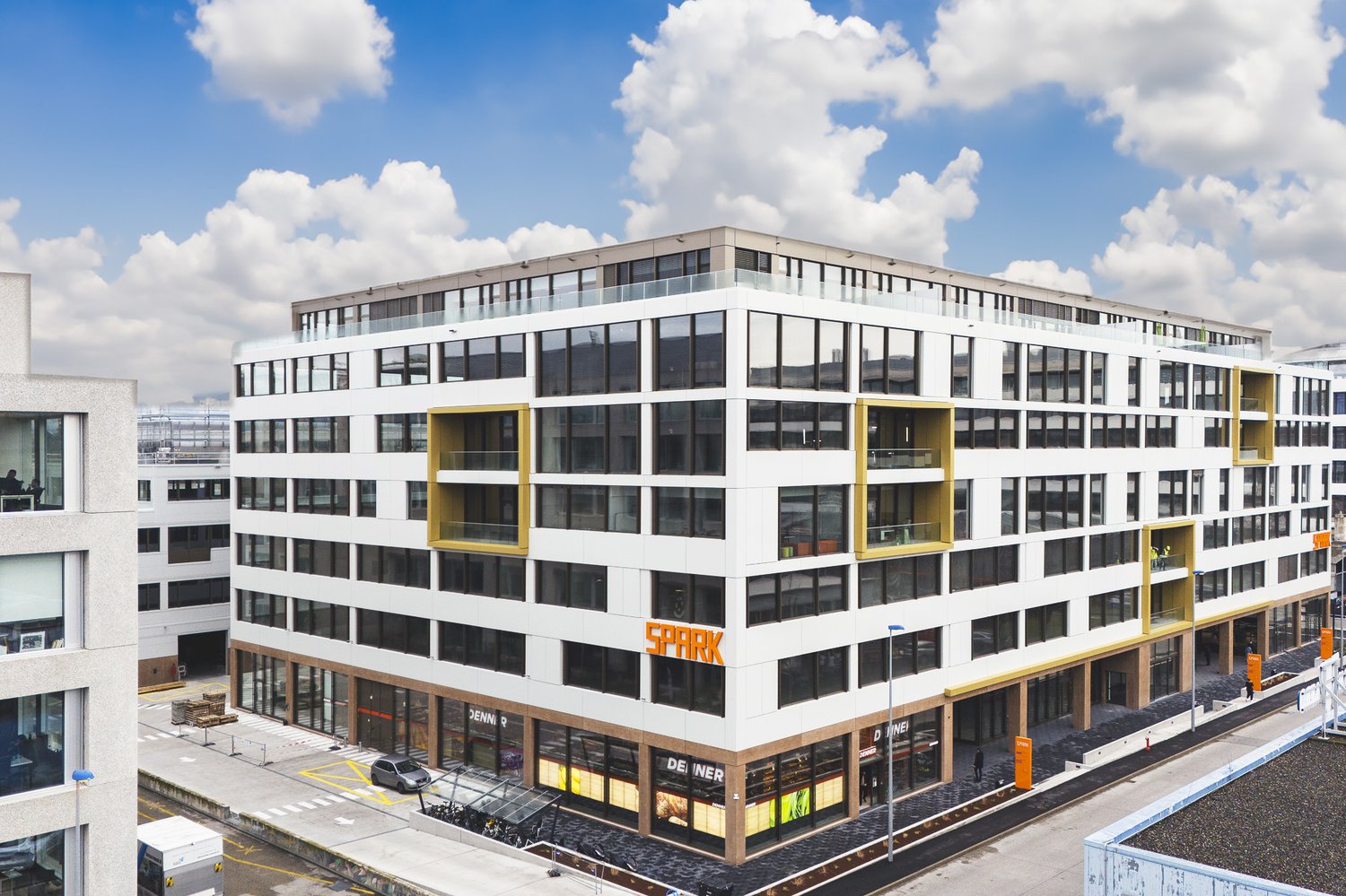 A modern, multi-story commercial building with a large glass facade, yellow accents, and a parking area in front.
