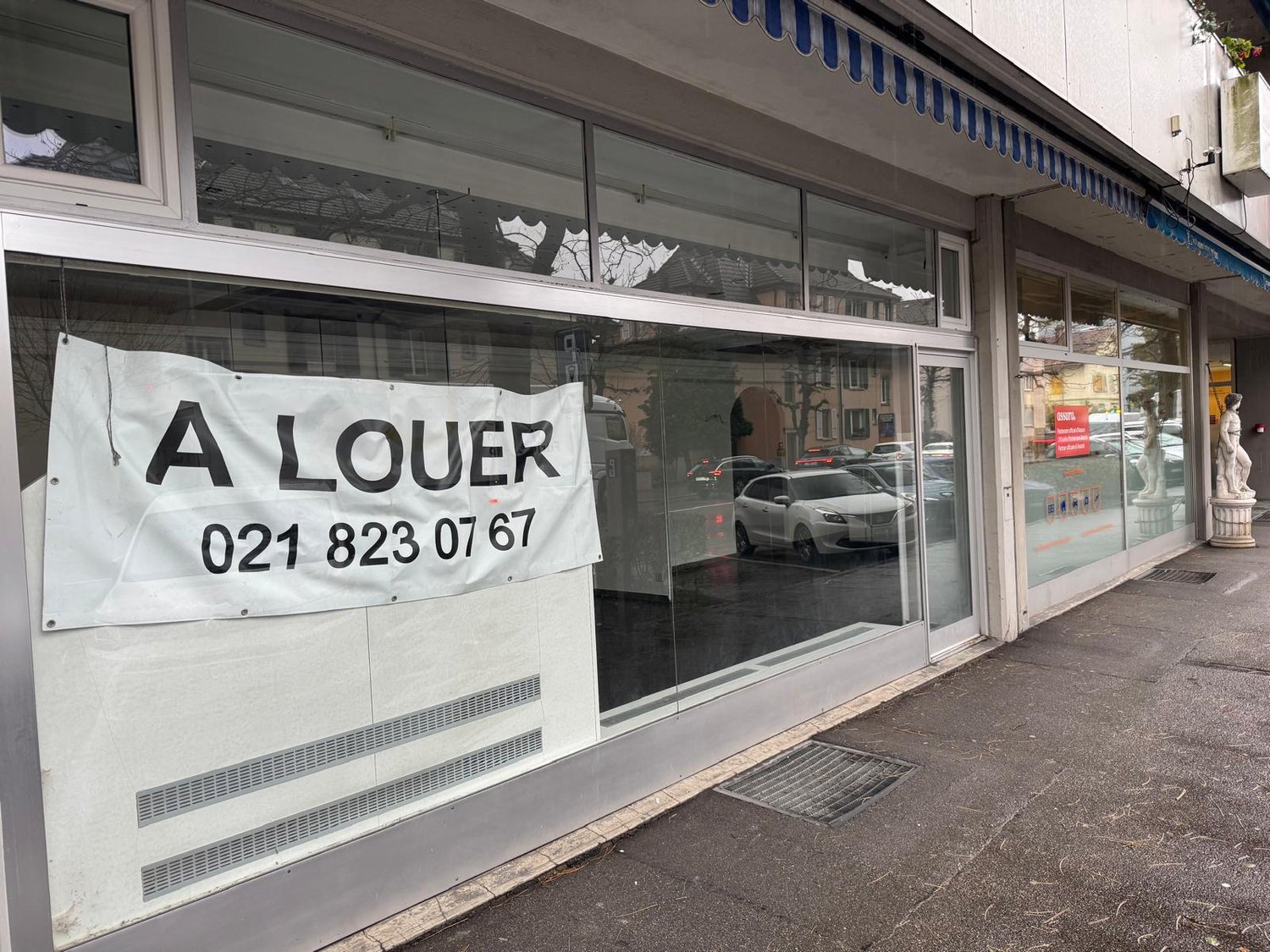 Shopfront with glass walls and doors, blue awning, white exterior, parked cars outside, signboard with a phone number and 'A LOUER' written in black