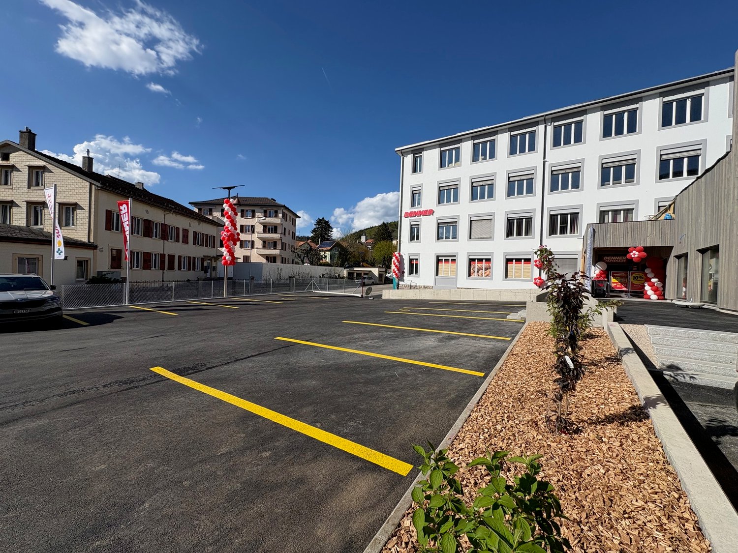 A multi-story commercial building with a large parking lot in front. The building has a modern, white facade with numerous windows. The parking lot is marked with yellow lines and contains several parked cars. There are some decorative plants and flowers