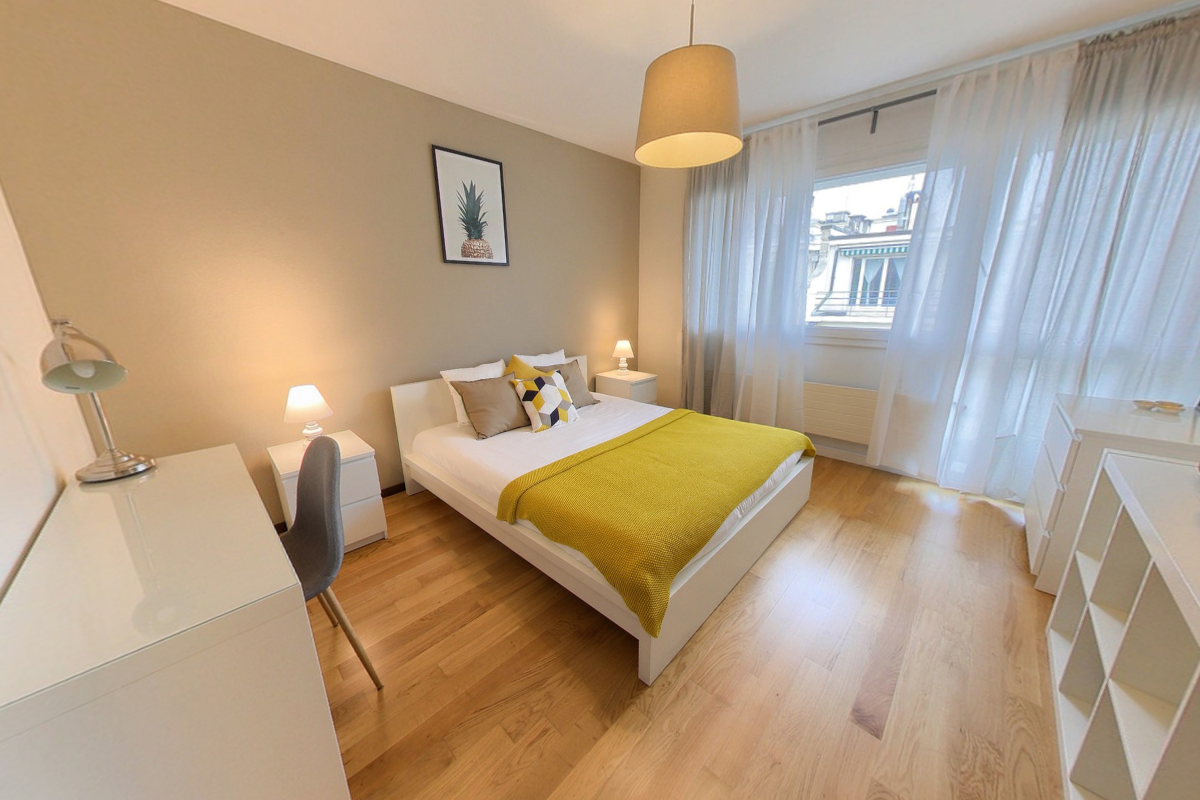 Spacious bedroom with hardwood flooring, white walls, large windows, a double bed with yellow bedding, nightstands, and a pendant light fixture.