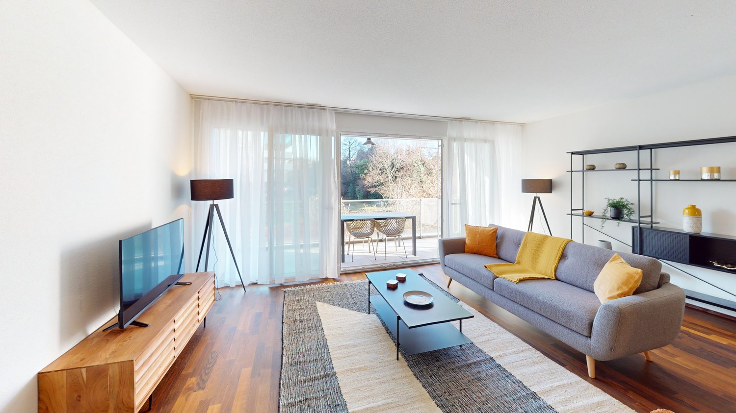 modern living room, gray sofa, wooden floor, sliding glass door to terrace, gray and yellow cushions, coffee table, TV, standing lamps, white curtains, decorative shelves