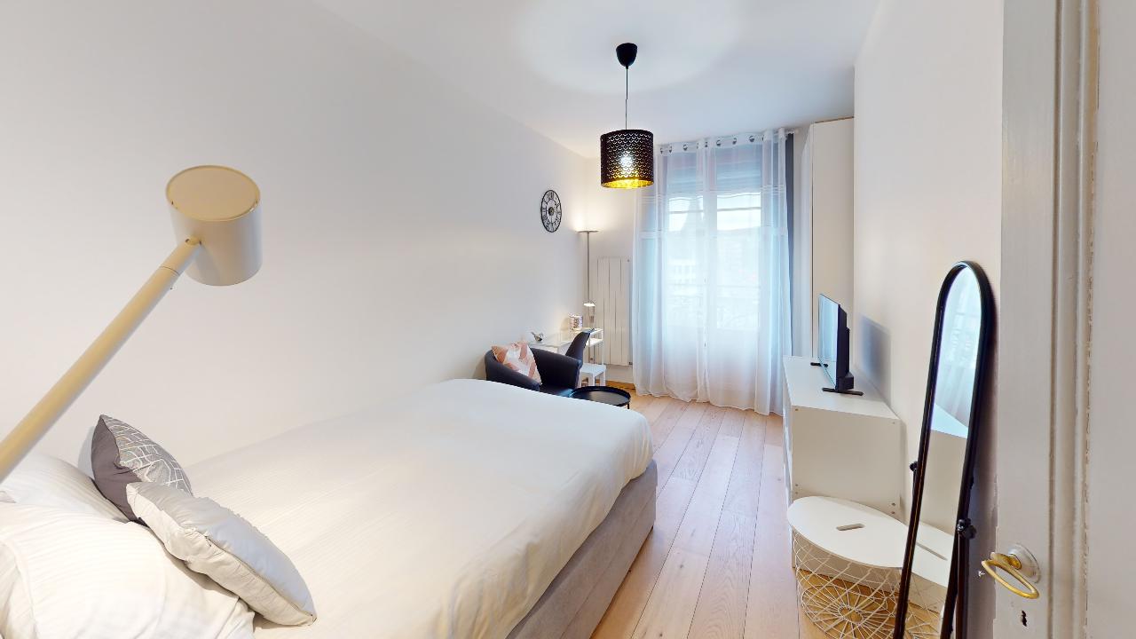 Spacious bedroom with white walls, hardwood floors, large window, pendant light fixture, and decorative accents.