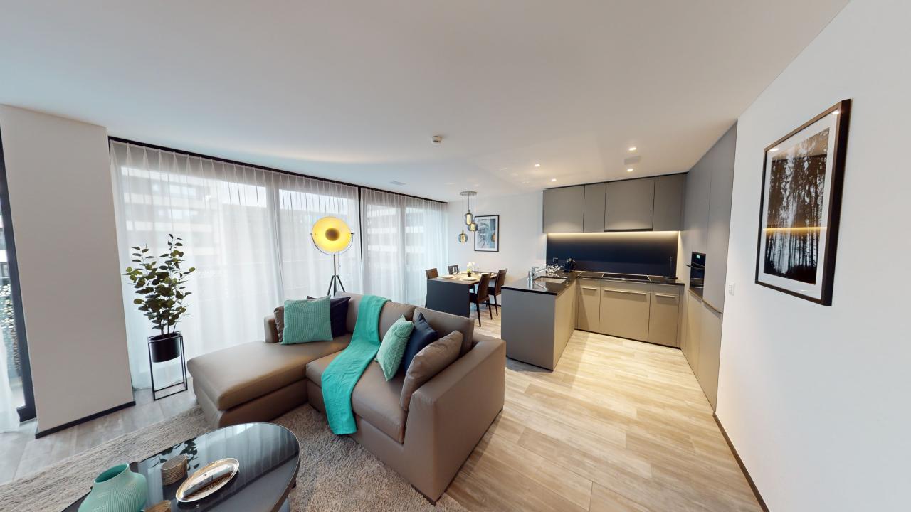 Open-concept living room and kitchen, modern decor with gray sofa, turquoise accent pillows, large windows, hardwood flooring, kitchen with dark cabinets and stainless steel appliances
