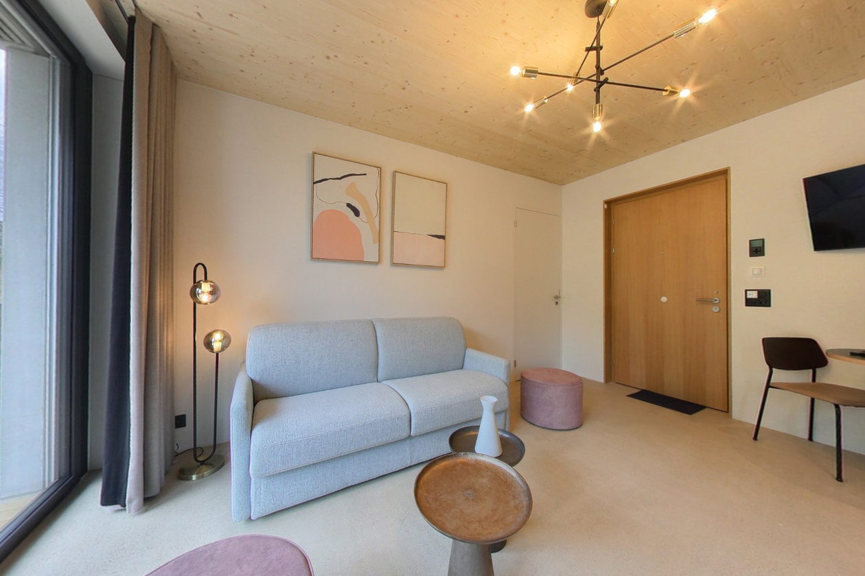 Small living room with light gray sofa, small tables, floor lamp, rug, chair, ottoman, television mounted on wall, paintings on wall