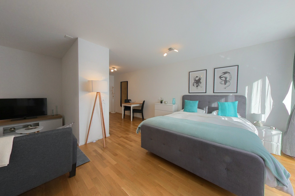 Bedroom with wooden floor, white walls, bed with blue pillows, two nightstands, bedside lamps, wall-mounted pictures, a table and chairs in a corner, a TV, a lamp, and a chair.