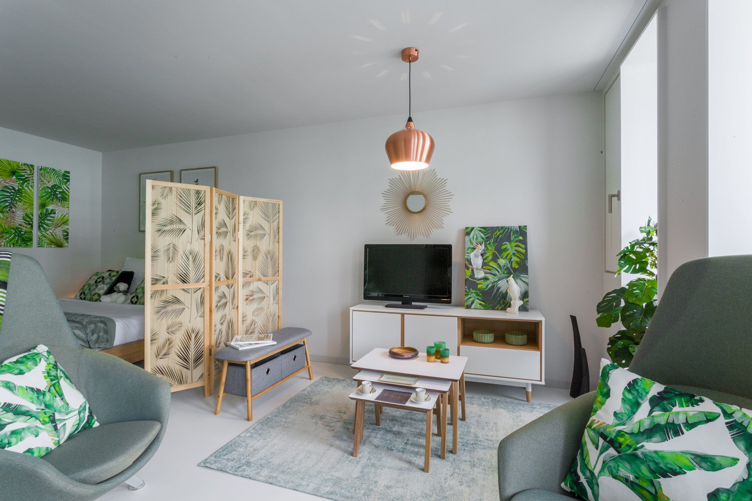 Bright and airy living room with white walls, hardwood floors, and tropical-inspired decor. Features a copper pendant light, a wooden screen divider, a TV stand with storage, and a seating area with a gray sofa and armchair. The room also has potted plant