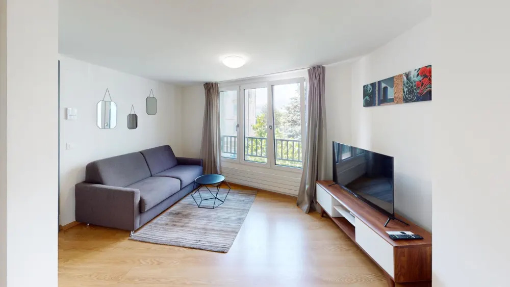 Spacious living room with large windows, hardwood floors, gray sofa, coffee table, and wall-mounted TV. The room has a balcony or terrace visible through the windows.