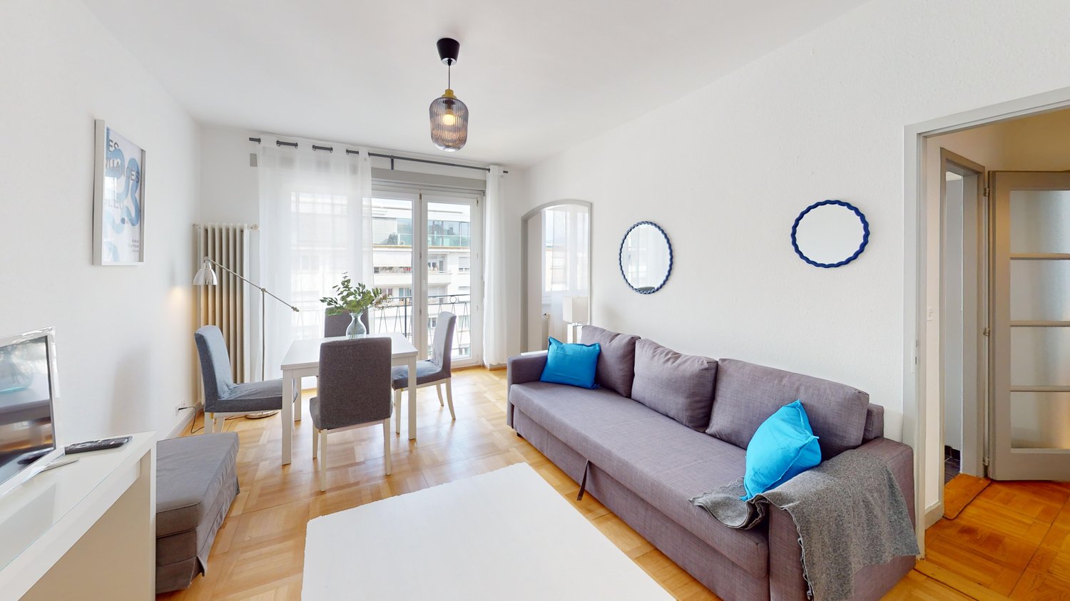 living room with dining area, gray couch, white table, chairs, white rug, white walls, wooden floor, white curtains, lamps, mirror, picture frame, television
