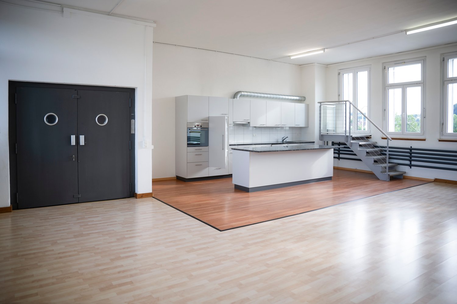 Spacious open-plan kitchen with white cabinets, built-in appliances, and hardwood flooring. Large windows provide natural light. Metal staircase visible in the background.