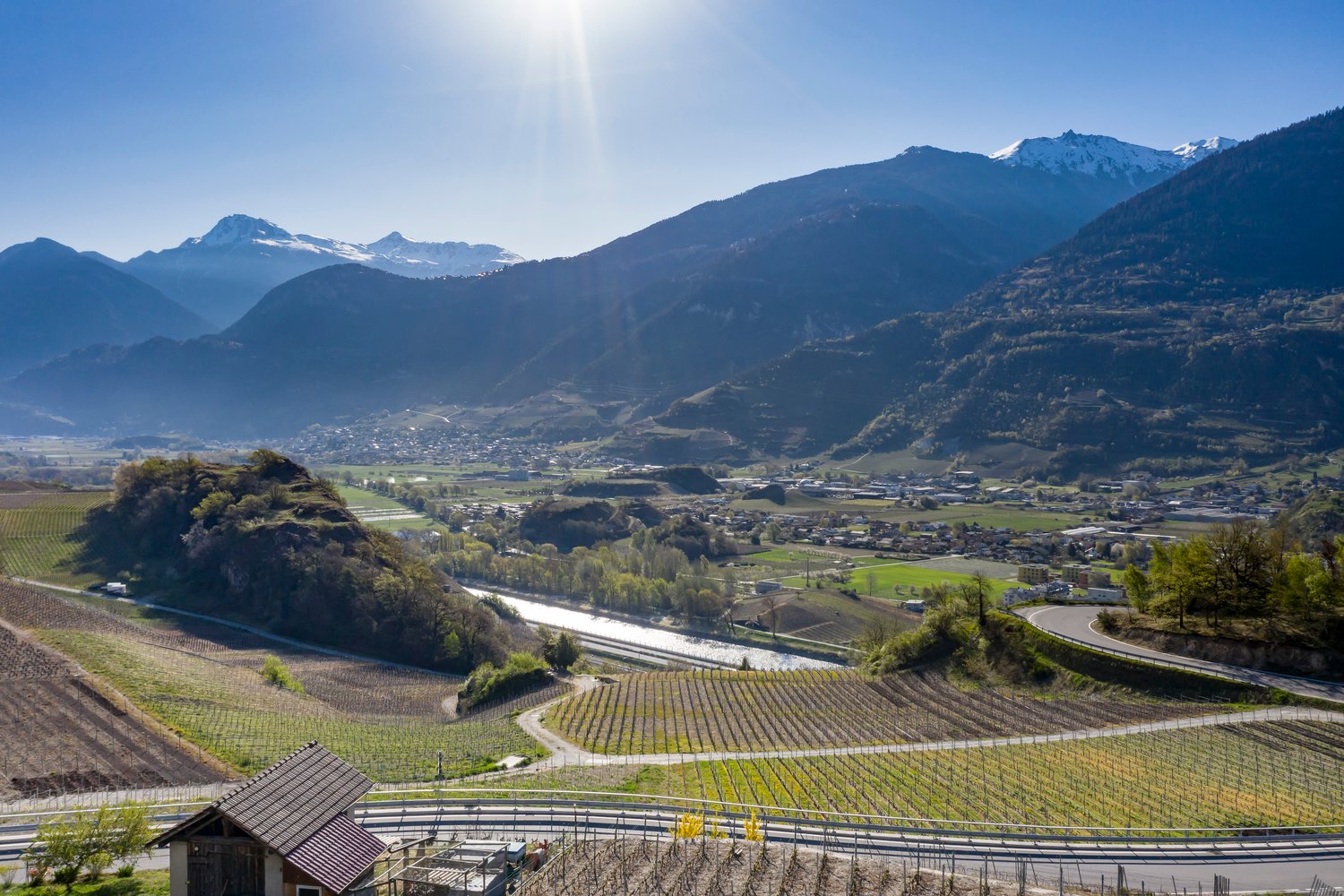The image shows a picturesque mountain landscape with snow-capped peaks in the background. In the foreground, there are rolling hills covered in vineyards and farmland, with a small town nestled in the valley below. The scene is bathed in warm, golden sun