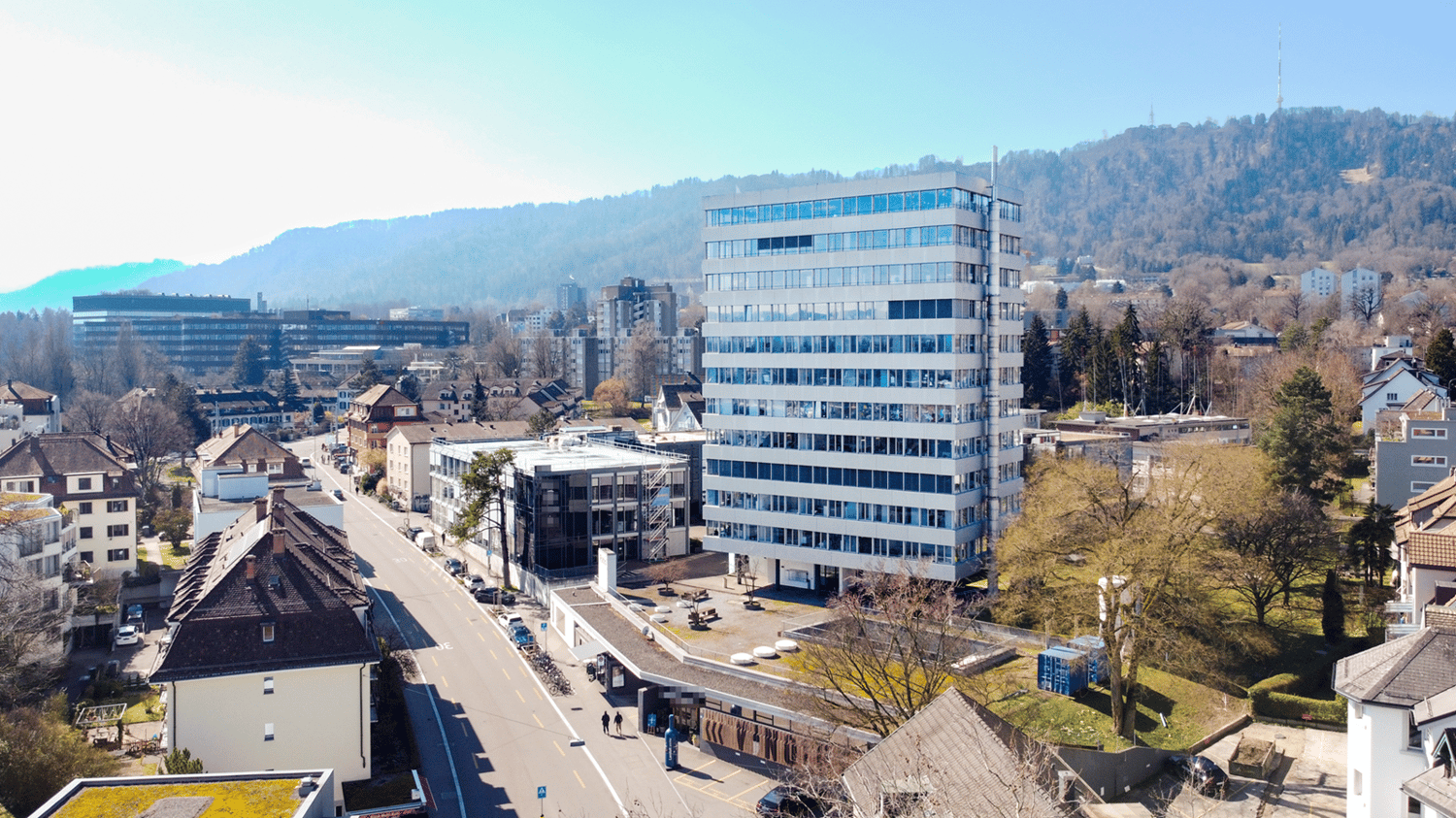 Office building with many windows, surrounded by other buildings, trees, and mountains in the background