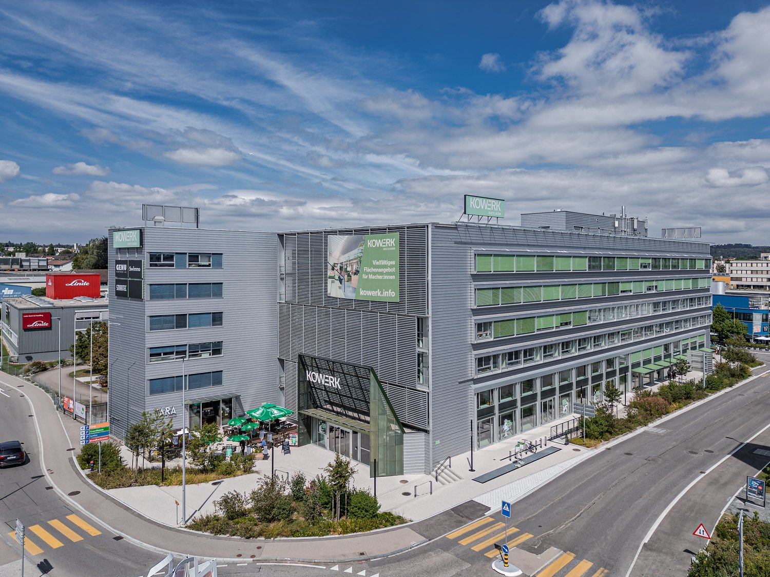Large multi-story commercial building with the Kowerk logo and branding. The building has a modern, glass and metal facade with several floors. There are parking spaces visible in the foreground of the image.