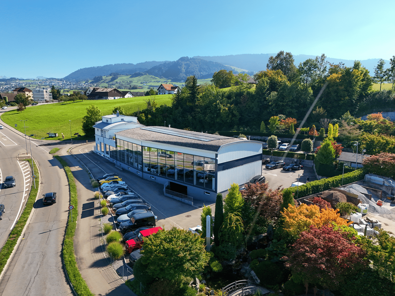 commercial building with multiple car parking spots, solar panels on the roof, large windows, modern design, trees, green field and hills in the background
