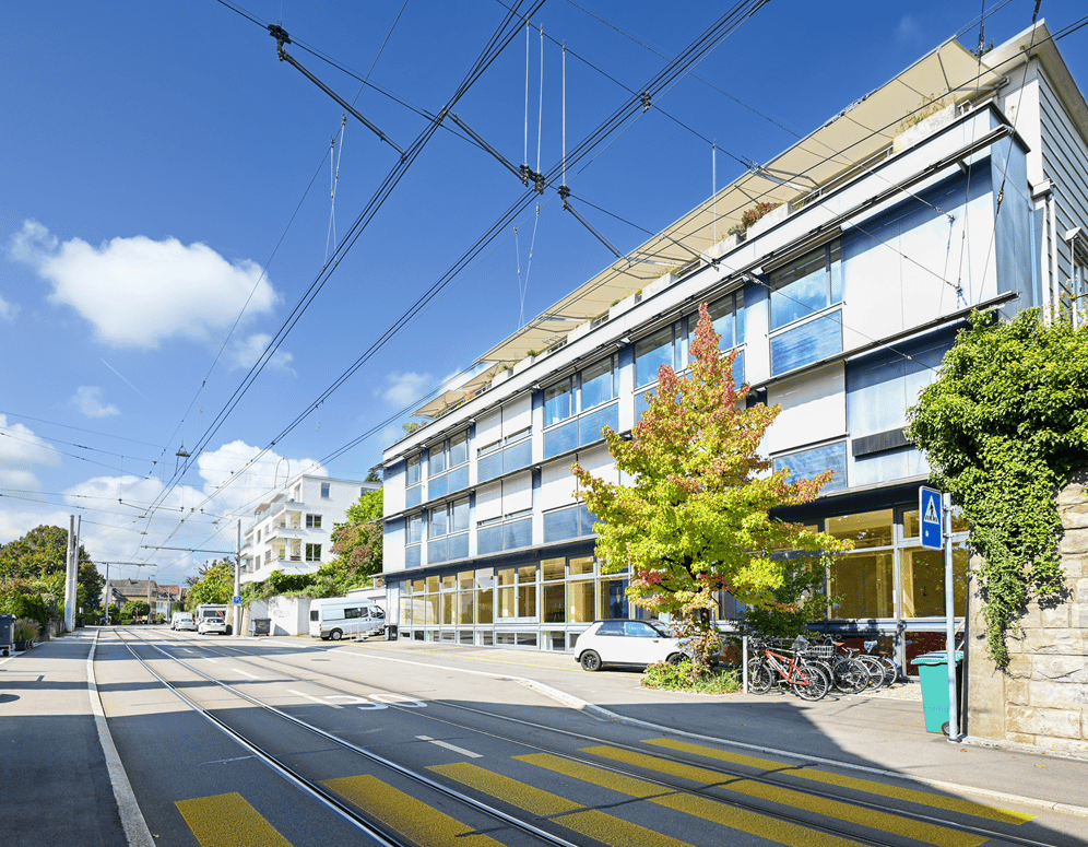 modern blue and white building, multiple floors, large glass windows, cars parked, bicycles leaning against wall, street light, greenery