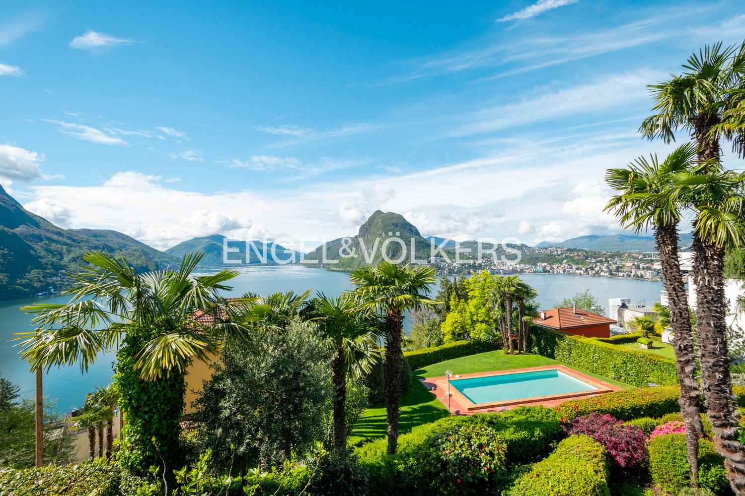 Luxury villa with swimming pool, surrounded by lush tropical gardens and palm trees, overlooking a scenic lake and mountains in the background.