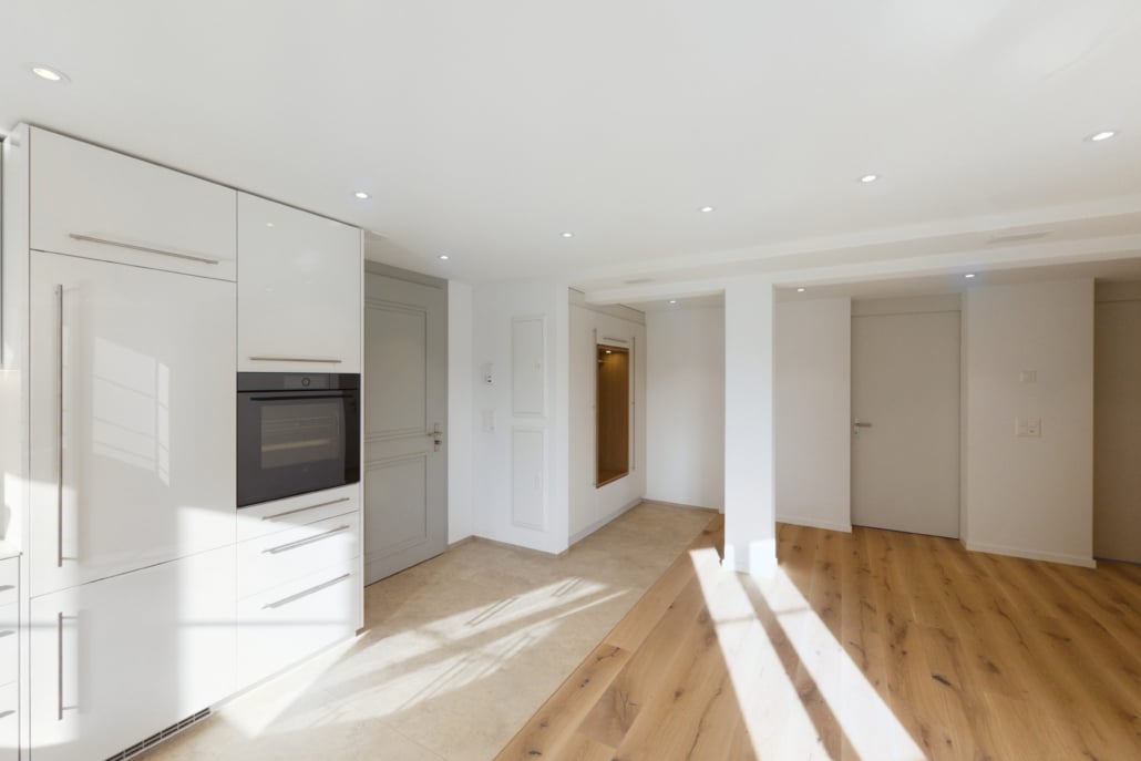 Open-concept living space with white walls, hardwood floors, built-in kitchen with oven and dishwasher, and a hallway leading to other rooms