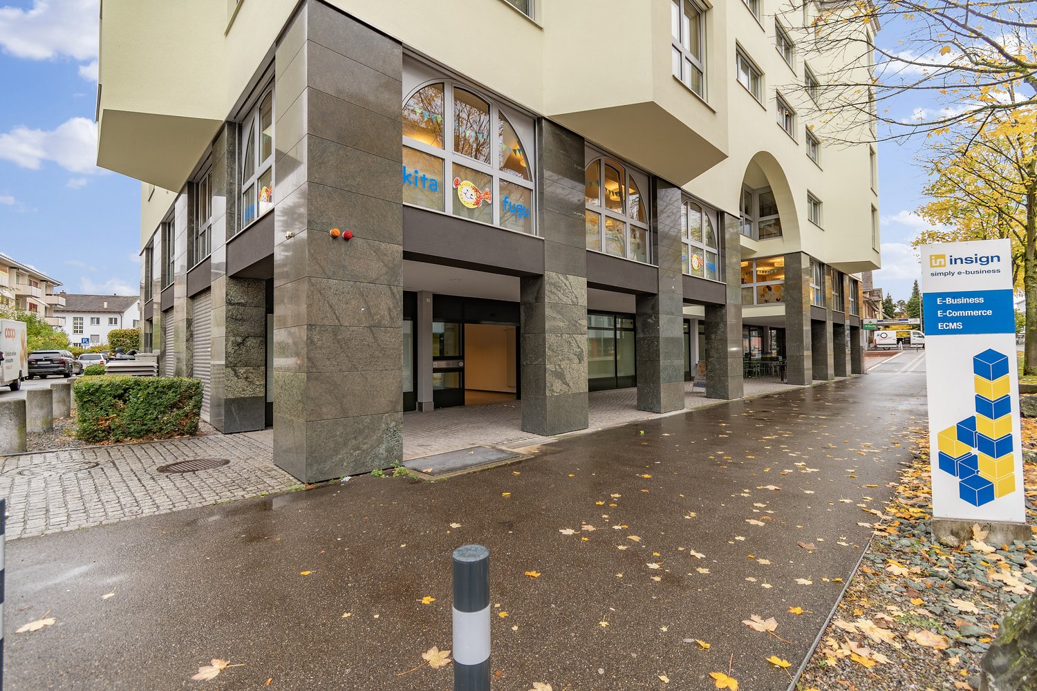 A multi-story commercial building with a modern, glass-fronted design. The building has a stone facade and a covered entrance. There are several businesses operating in the building, including Kita and Insign. The surrounding area has trees with autumn fo
