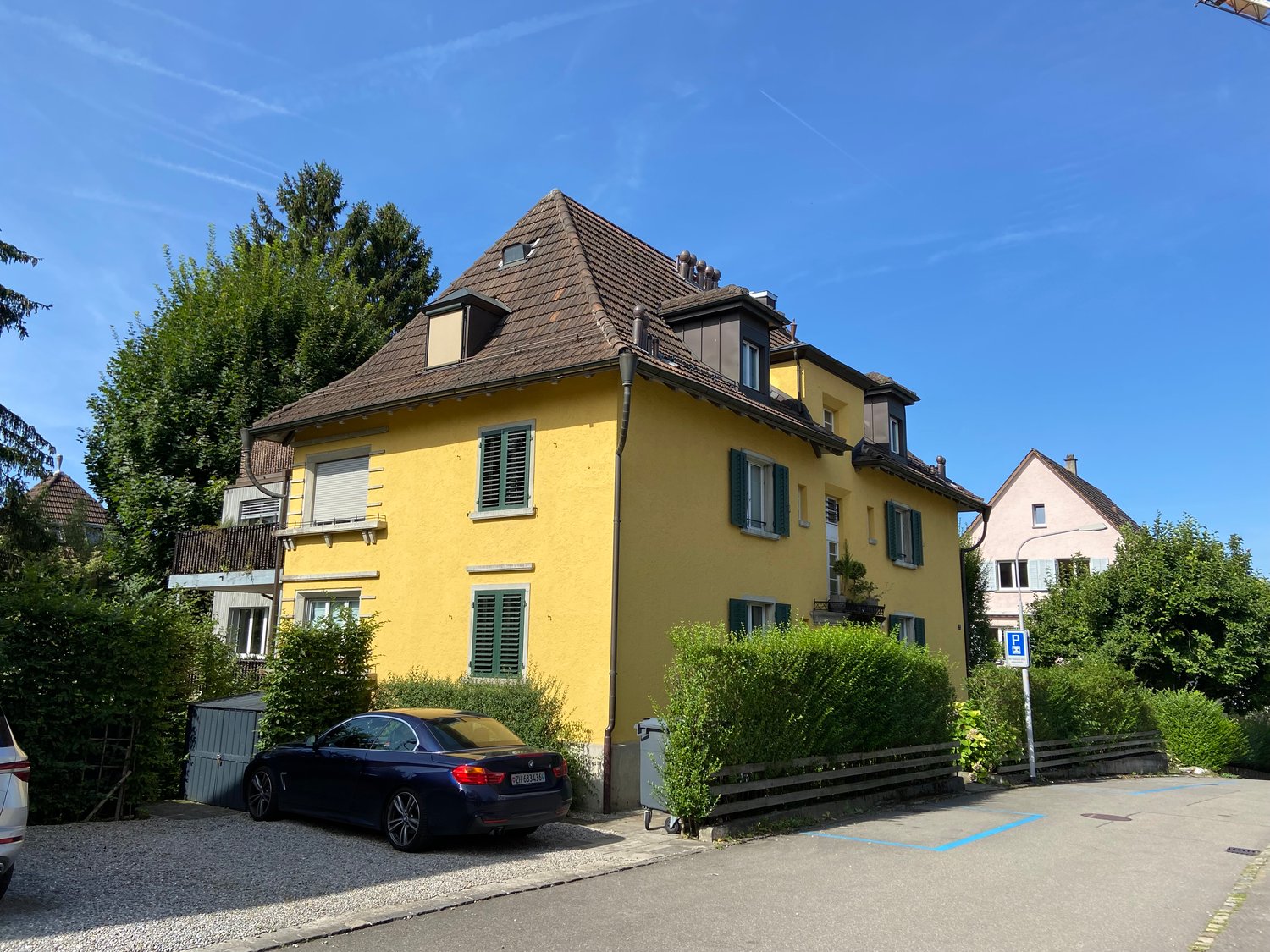 3-story yellow apartment building with a tiled roof, green shutters, and a parking space in front