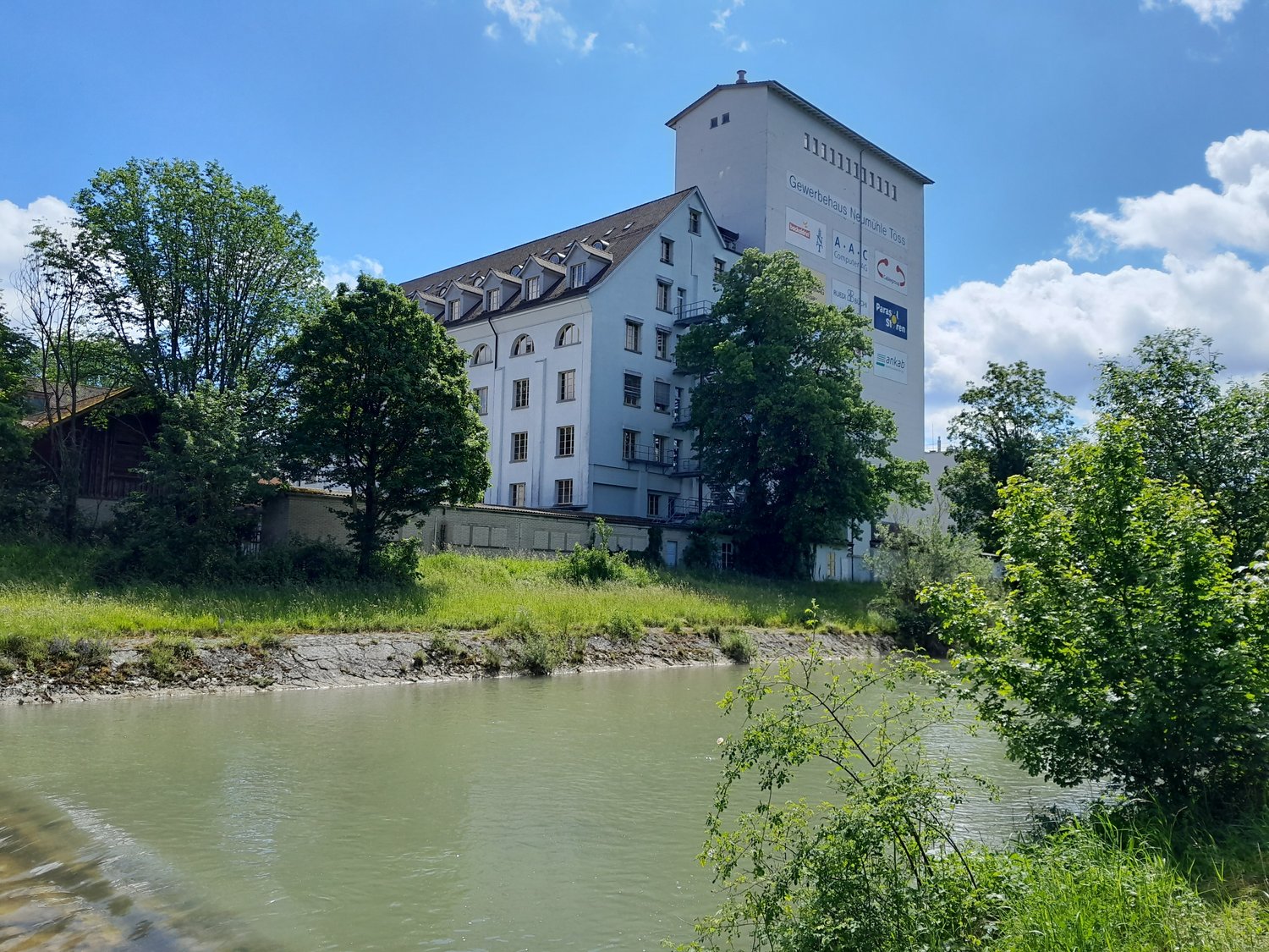 Large white building with multiple windows, situated next to a river, surrounded by trees and grass.
