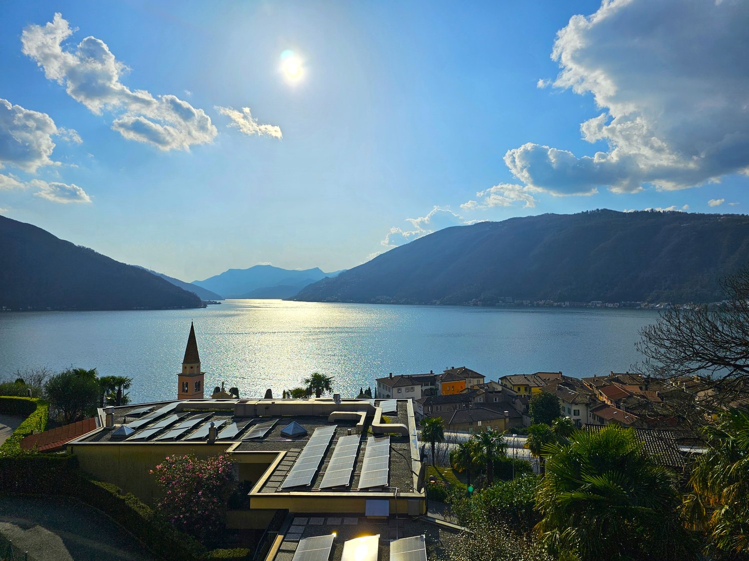 The image shows a scenic view of a lake surrounded by mountains. The lake is reflecting the sunlight, creating a shimmering effect on the water. In the foreground, there is a town with colorful buildings and a church steeple visible. The landscape is lush