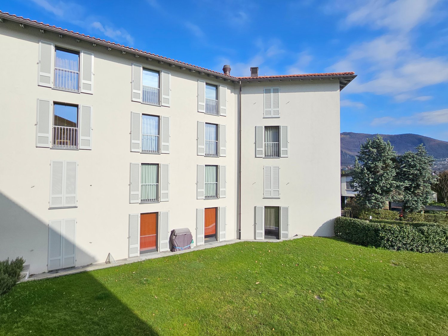 White building, several floors, windows with shutters, red doors, well-kept lawn, bushes, distant mountains