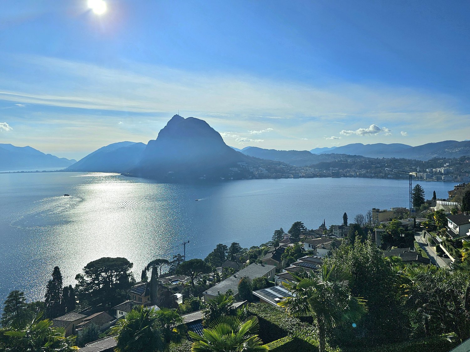 Lake Lugano with mountains in the background, a clear blue sky, town in the distance, greenery