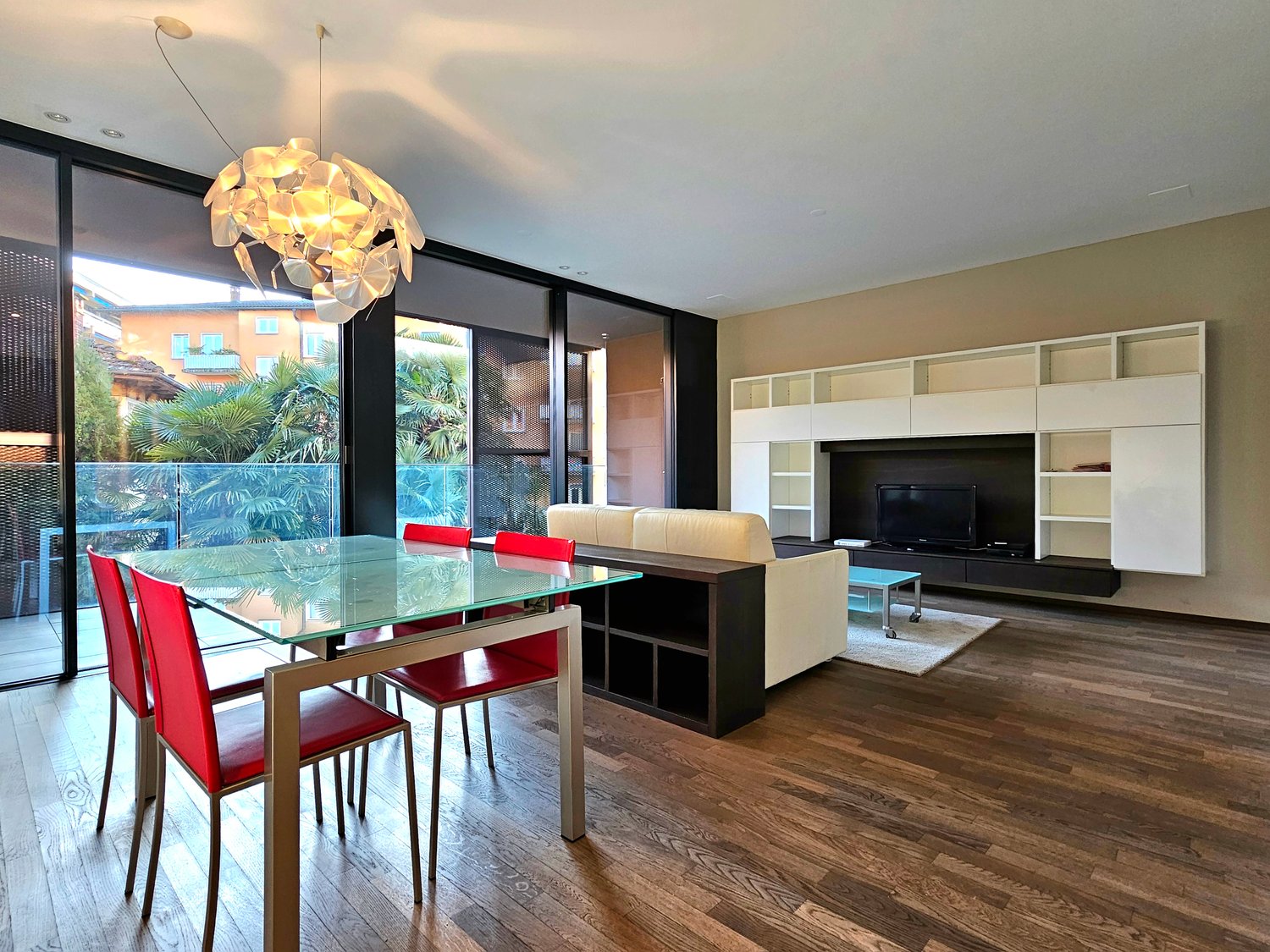 glass table, red chairs, white couch, TV, chandelier, wooden floor, glass walls