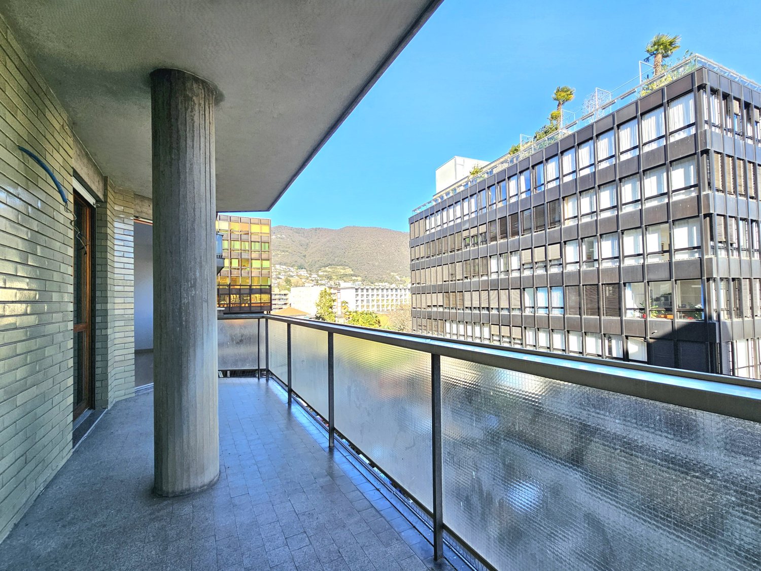 Balcony with glass railing, tiled floor, pillar, view of a building and mountain