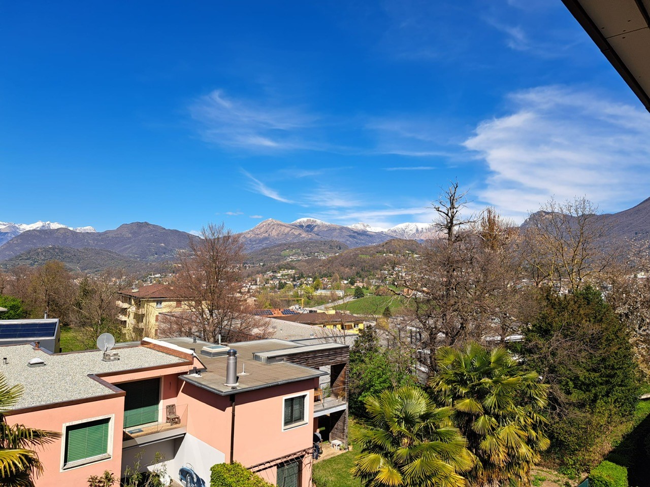Aerial view of a building with a pink facade, solar panels on the roof, and a balcony with a chair. The building is surrounded by trees and has a view of the mountains in the distance.