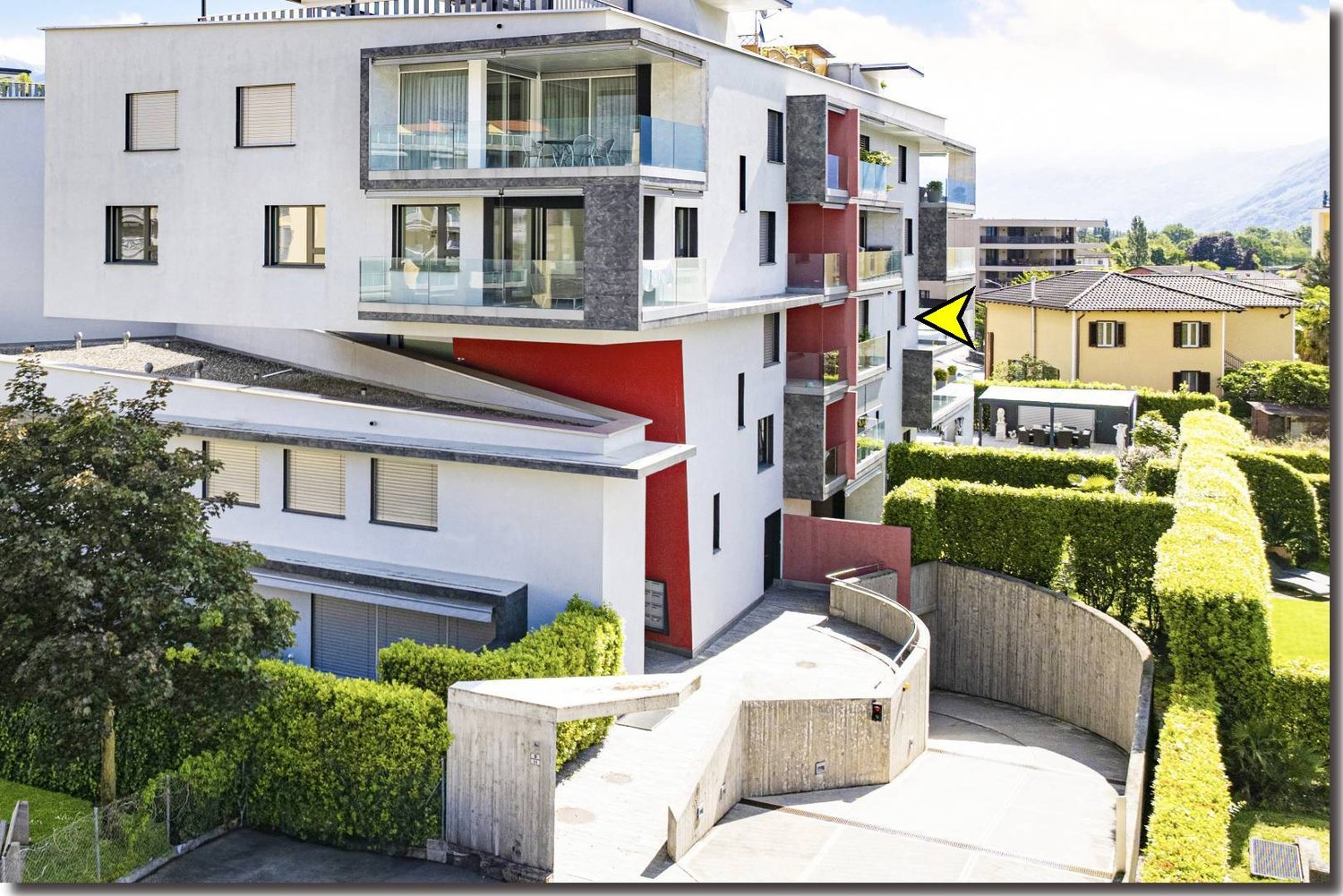 Modern apartment building with multiple floors, white and red facade, glass balconies, fenced entrance, hedges, and a small garden